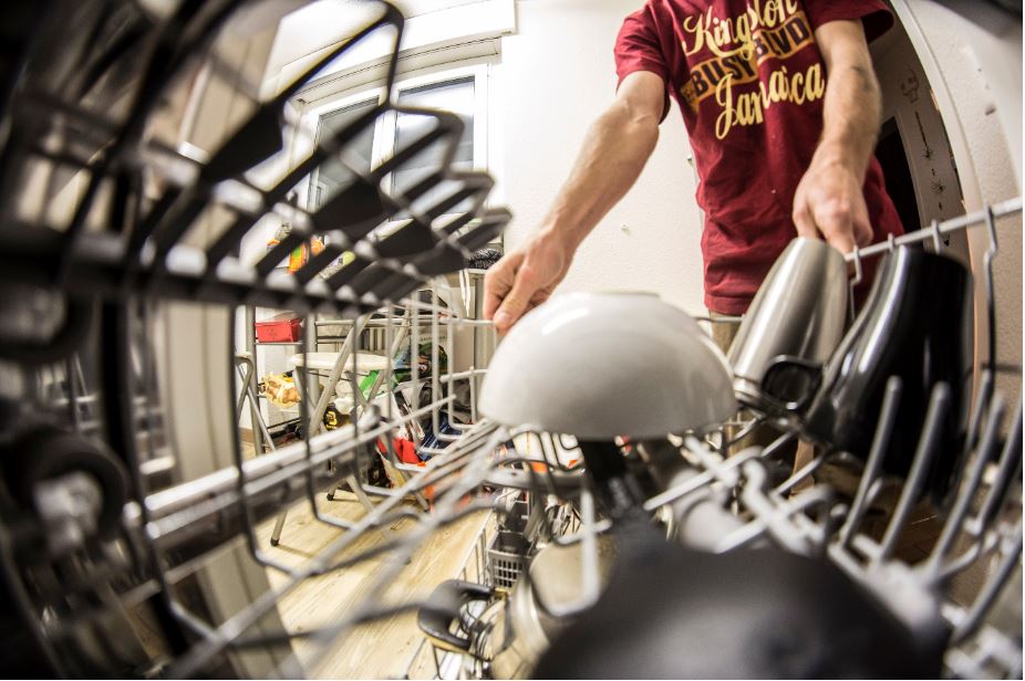 A man captured holding dish racks inside a dishwasher with a big white ceramic bow upright